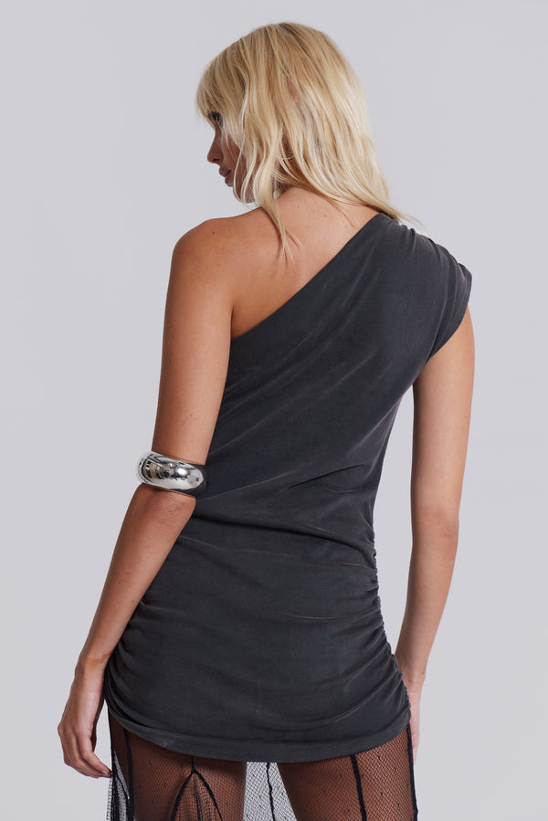 Female model wearing a grey tonal screenprint one shoulder ruched mini dress, styled with a black sheer mesh maxi length skirt. 