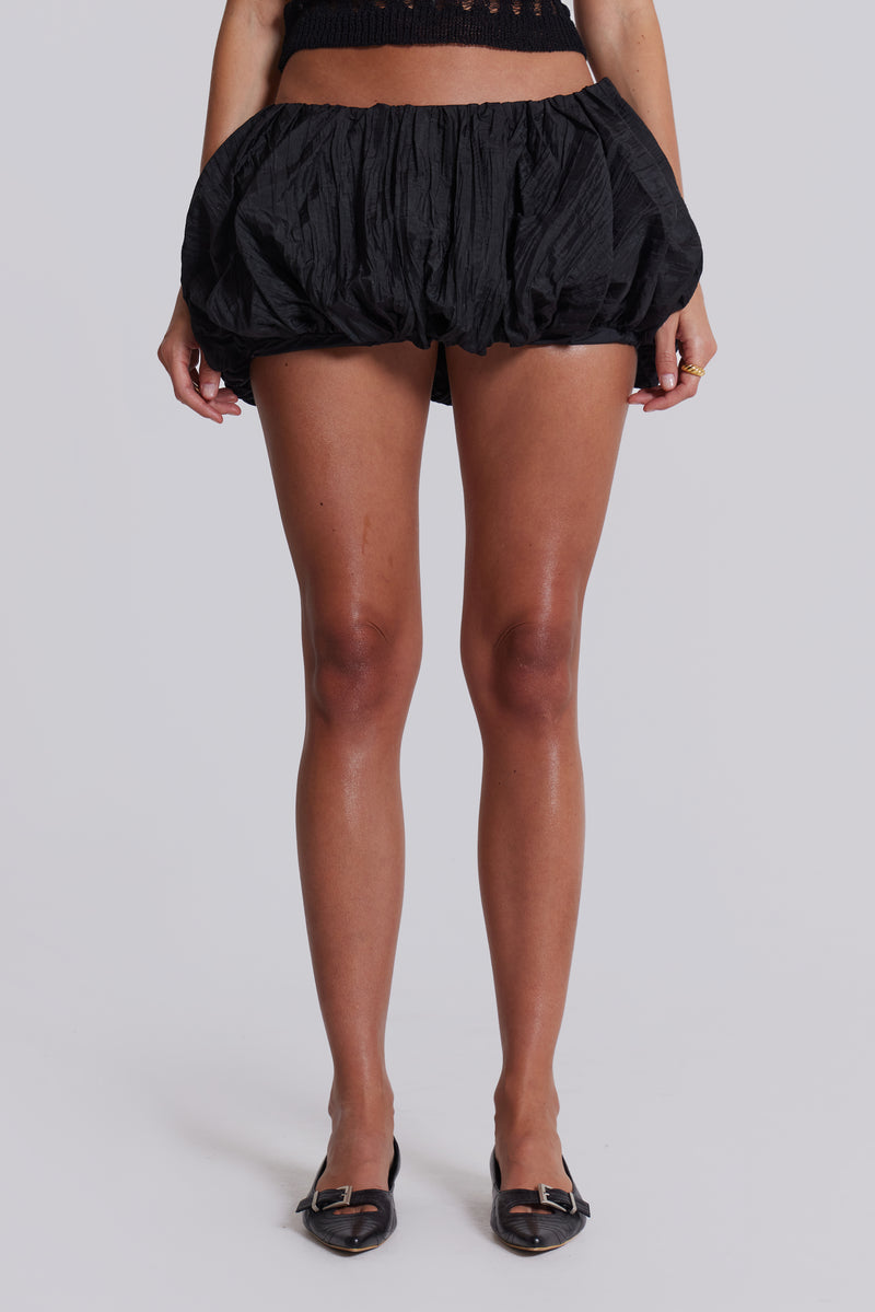 Female model wearing a black balloon style taffeta mini skirt with a puffball hem. Styled with a black knitted top. 