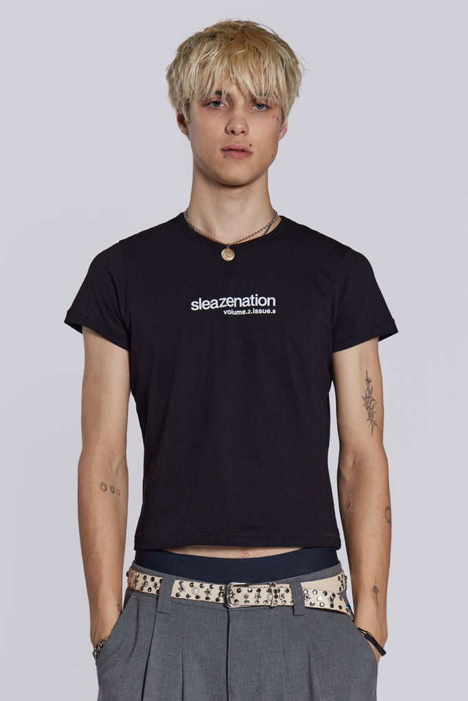 Male wearing black slogan t-shirt, styled with grey pleated trousers and studded belt.