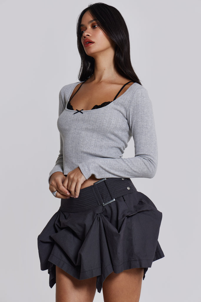 Female model wearing a grey marl long sleeve pointelle fabric with rib binding and needle neckline finish with a lace insert. Styled with a charcoal grey asymmetric tucked mini skirt. 