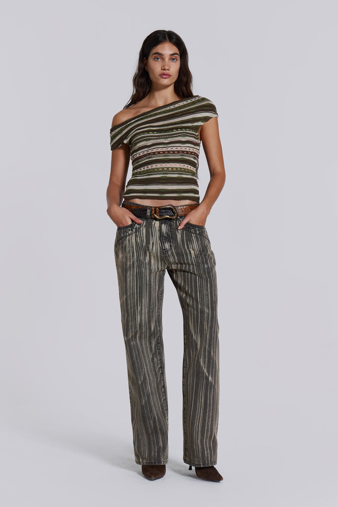  Female model wearing a tonal brown and khaki ruffle engineered hole knit fabric top with an asymmetric neckline. Styled with tonal striped jeans.