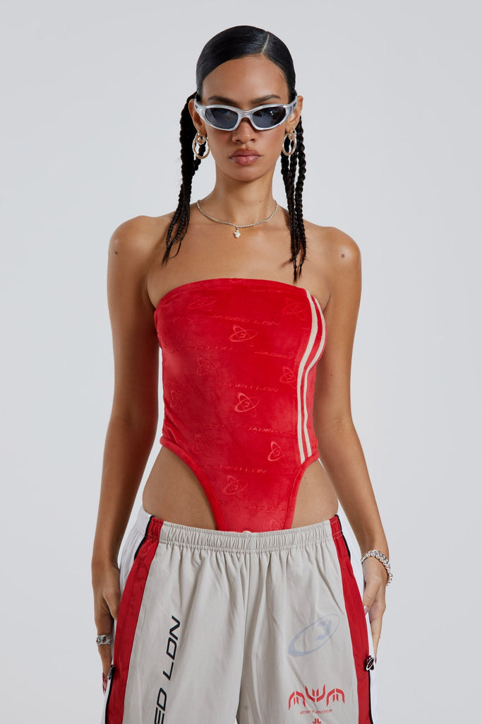 Female wearing red logo monogram embossed strapless bodysuit with stripe detail. Styled with almond track pants. 
