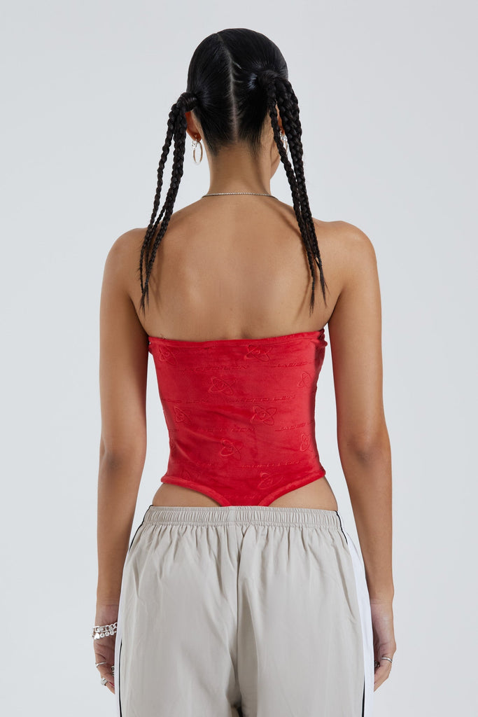 Female wearing red logo monogram embossed strapless bodysuit with stripe detail. Styled with almond track pants. 