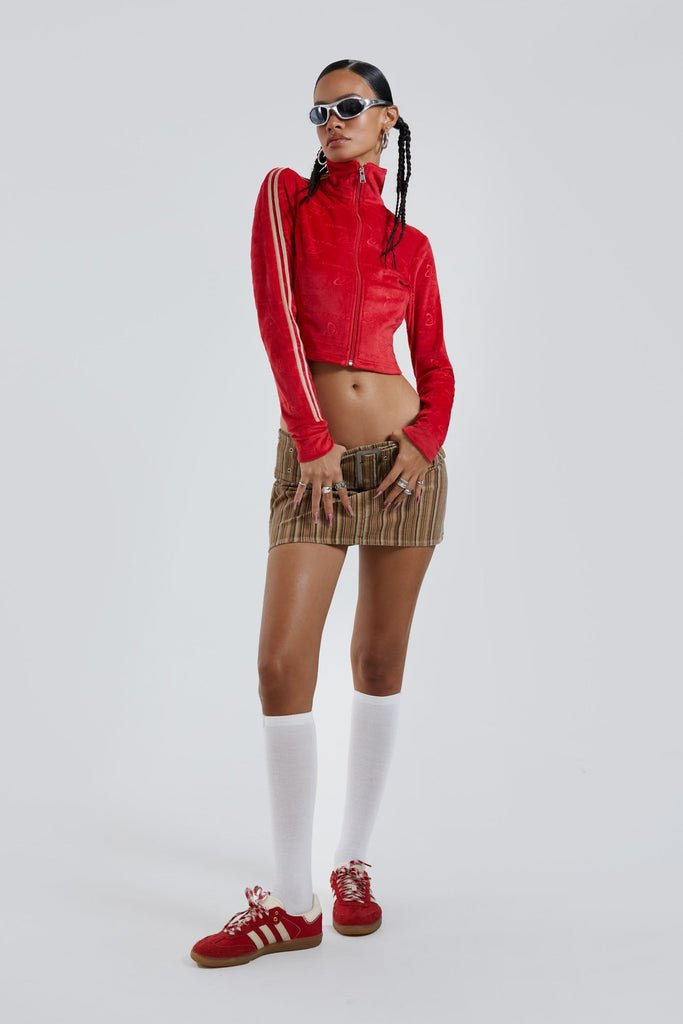 Female wearing red logo monogram embossed high neck long sleeve velour track top. Styled with brown stripe cord mini skirt. 