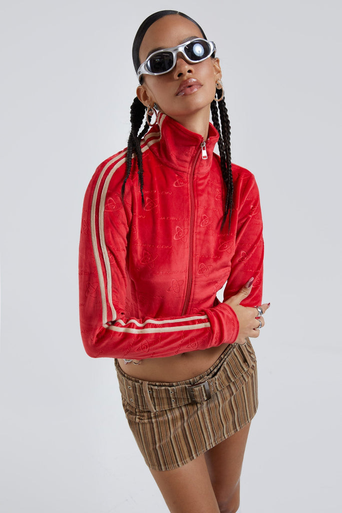 Female wearing red logo monogram embossed high neck long sleeve velour track top. Styled with brown stripe cord mini skirt. 