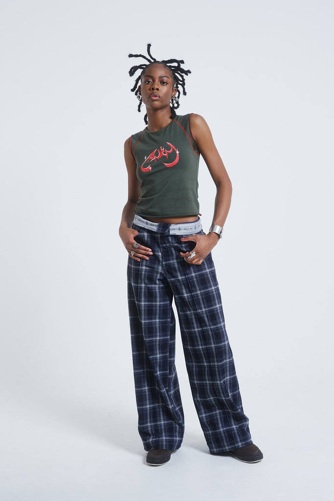 Female model wearing wide leg blue and white checked trousers with a fold over waist detail. Paired with a khaki green sleeveless vest top. 