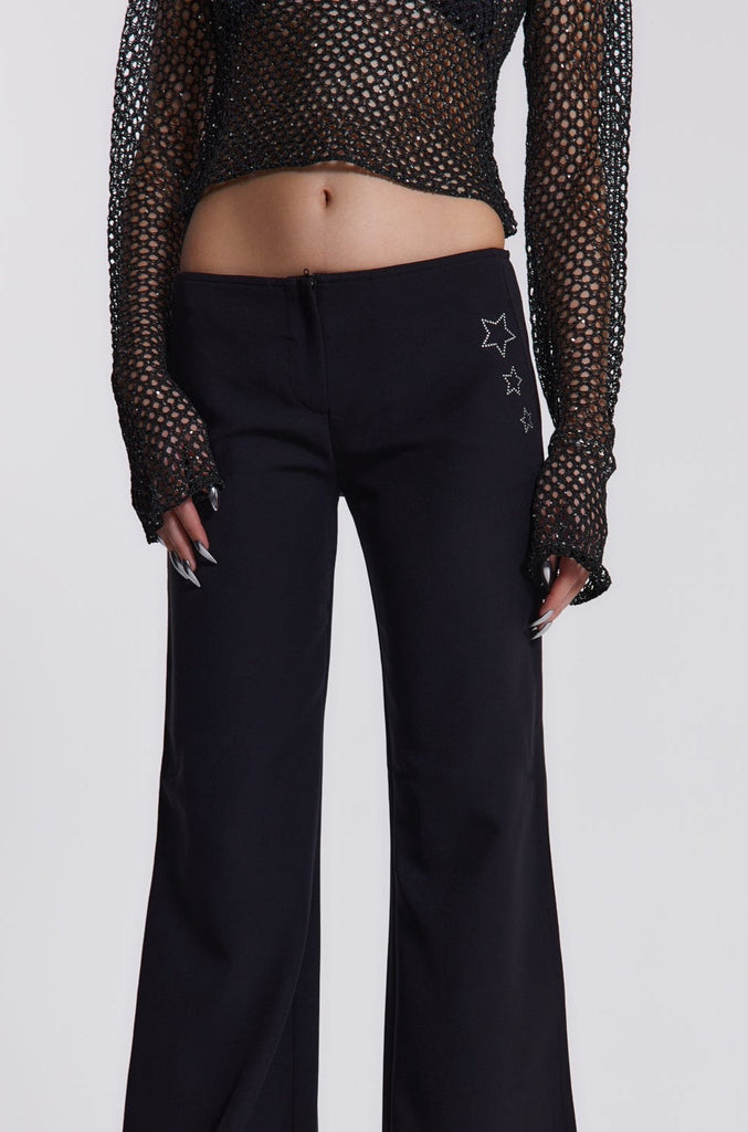 Female model wearing These black low waist trousers are in a wide leg fit and feature a silver diamante star detail.  Paired with a black crochet long sleeve top. 