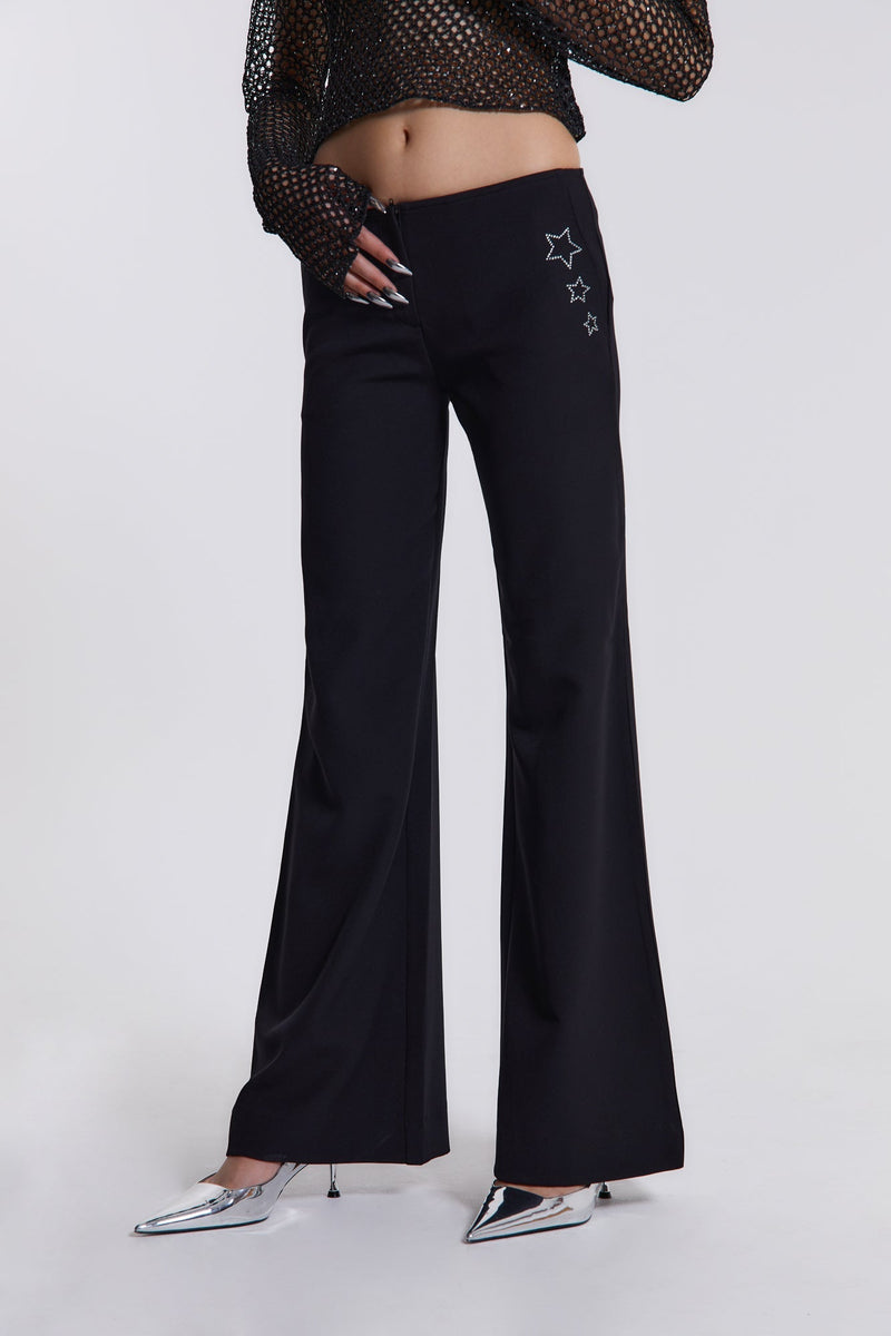 Female model wearing These black low waist trousers are in a wide leg fit and feature a silver diamante star detail.  Paired with a black crochet long sleeve top. 